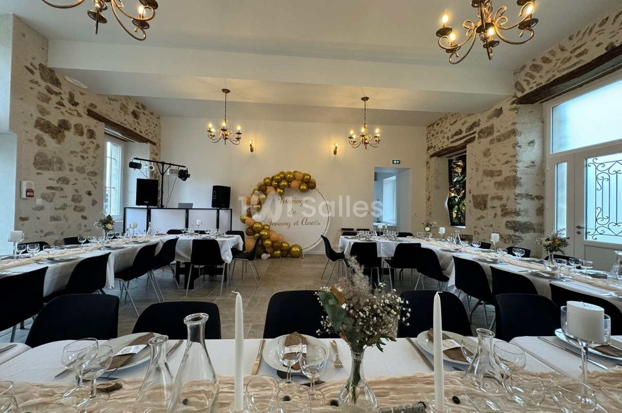 Salle de réception décorée pour un mariage, avec tables dressées, chandeliers au plafond et mur orné de ballons dorés.