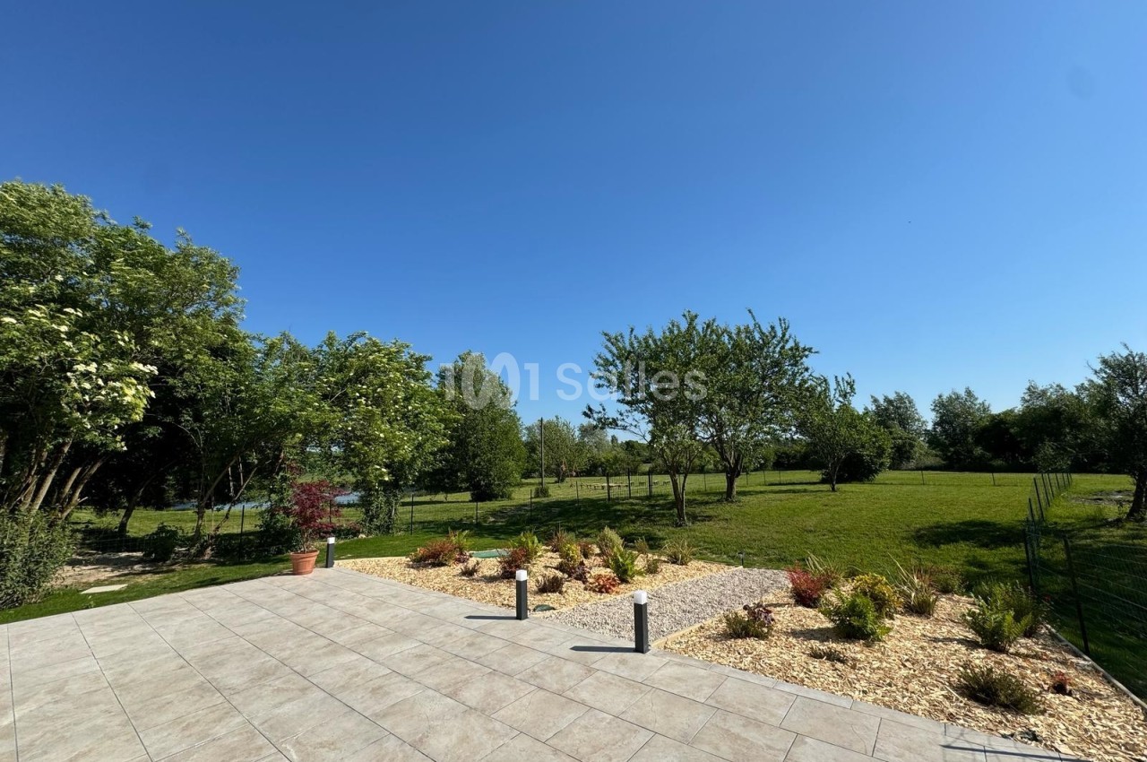 Vue d'une terrasse carrelée donnant sur un jardin arboré et une pelouse sous un ciel bleu dégagé.