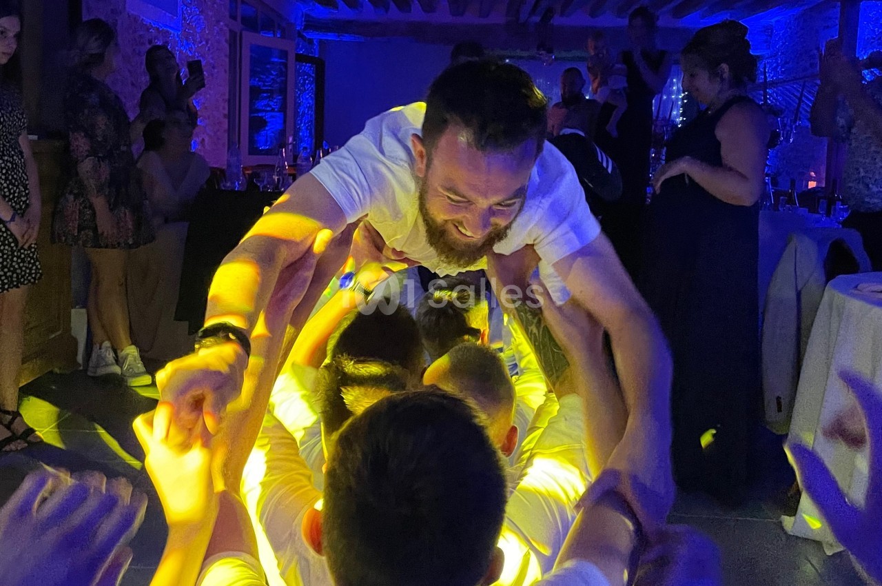 Un homme en chemise blanche porté par un groupe de personnes dans une ambiance festive et éclairage coloré.