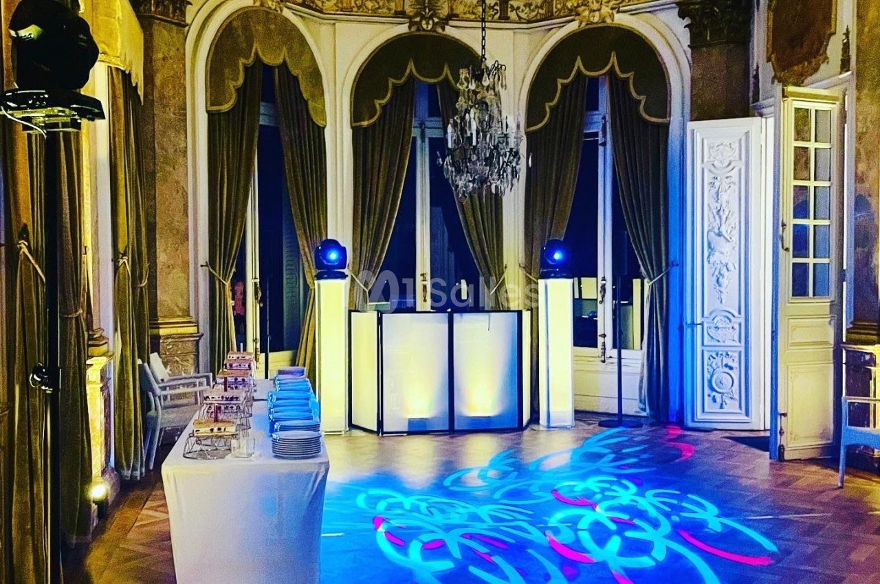 Salle élégante avec parquet, lustre, rideaux dorés, table dressée et éclairages colorés projetant des motifs au sol.