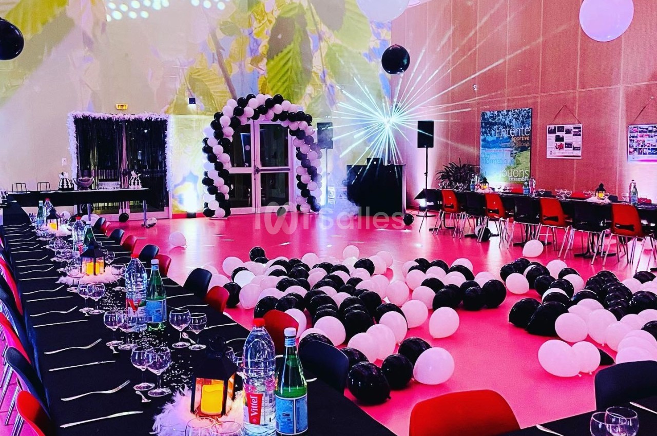 Salle décorée pour un événement avec tables dressées, ballons noirs et blancs, et éclairage coloré.