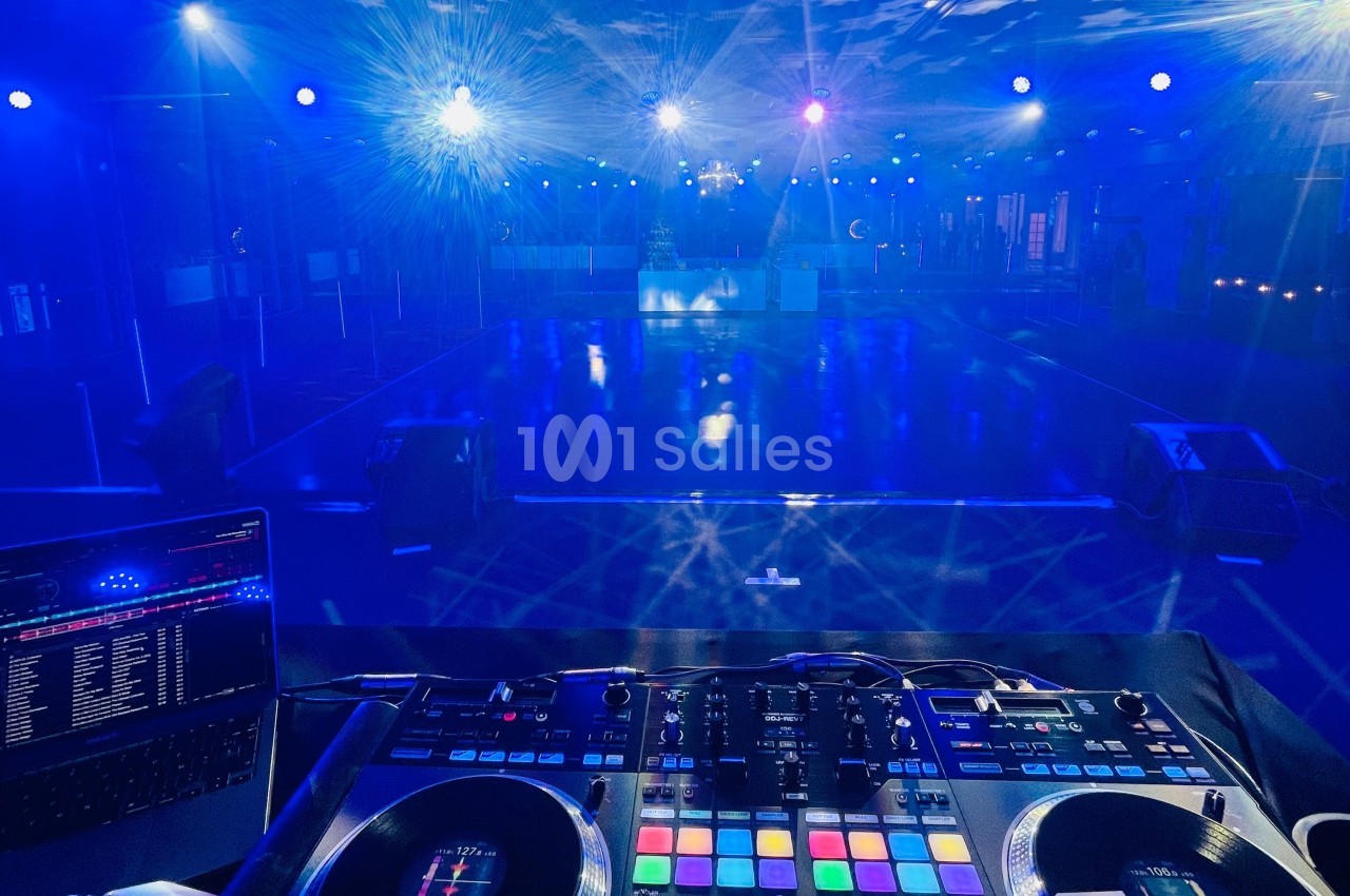 Vue depuis une table de mixage DJ éclairée, face à une piste de danse vide sous des lumières bleues.