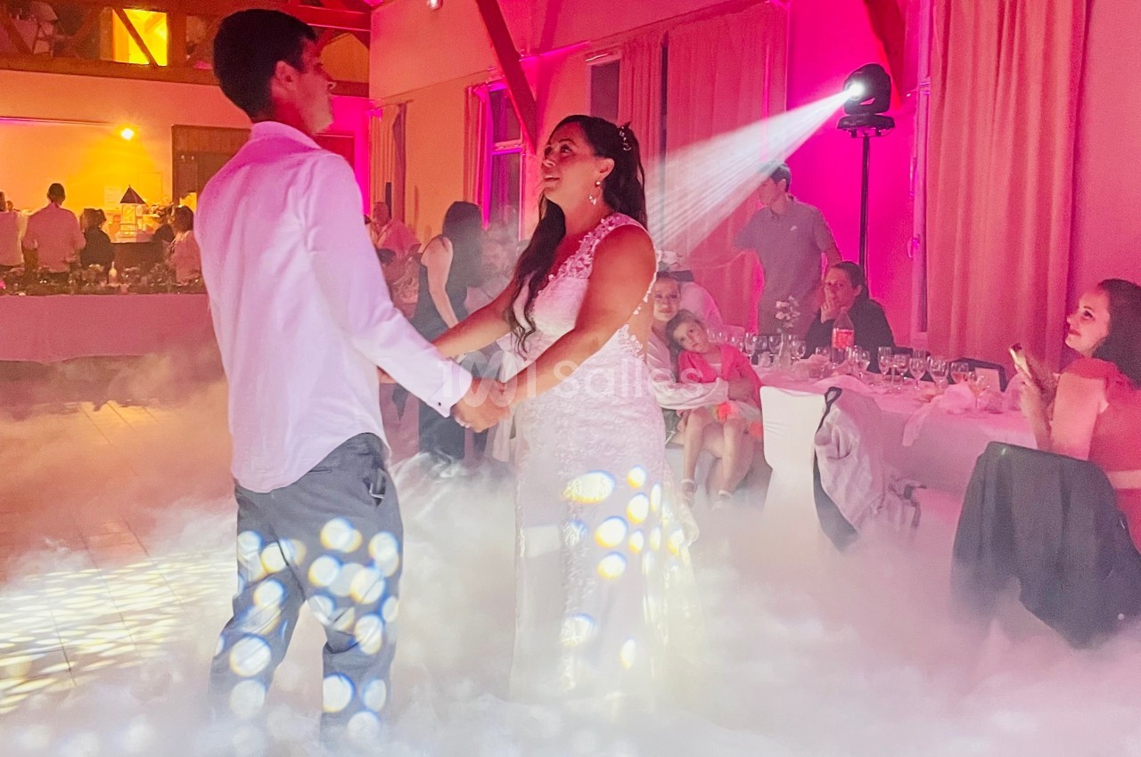 Un couple danse au milieu d'une piste entourée de fumée, sous des lumières colorées, devant des invités assis.