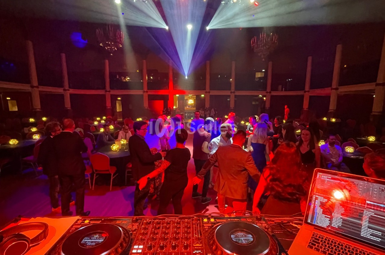 Vue depuis une table de mixage sur une piste de danse animée, éclairée par des projecteurs colorés dans une salle sombre.