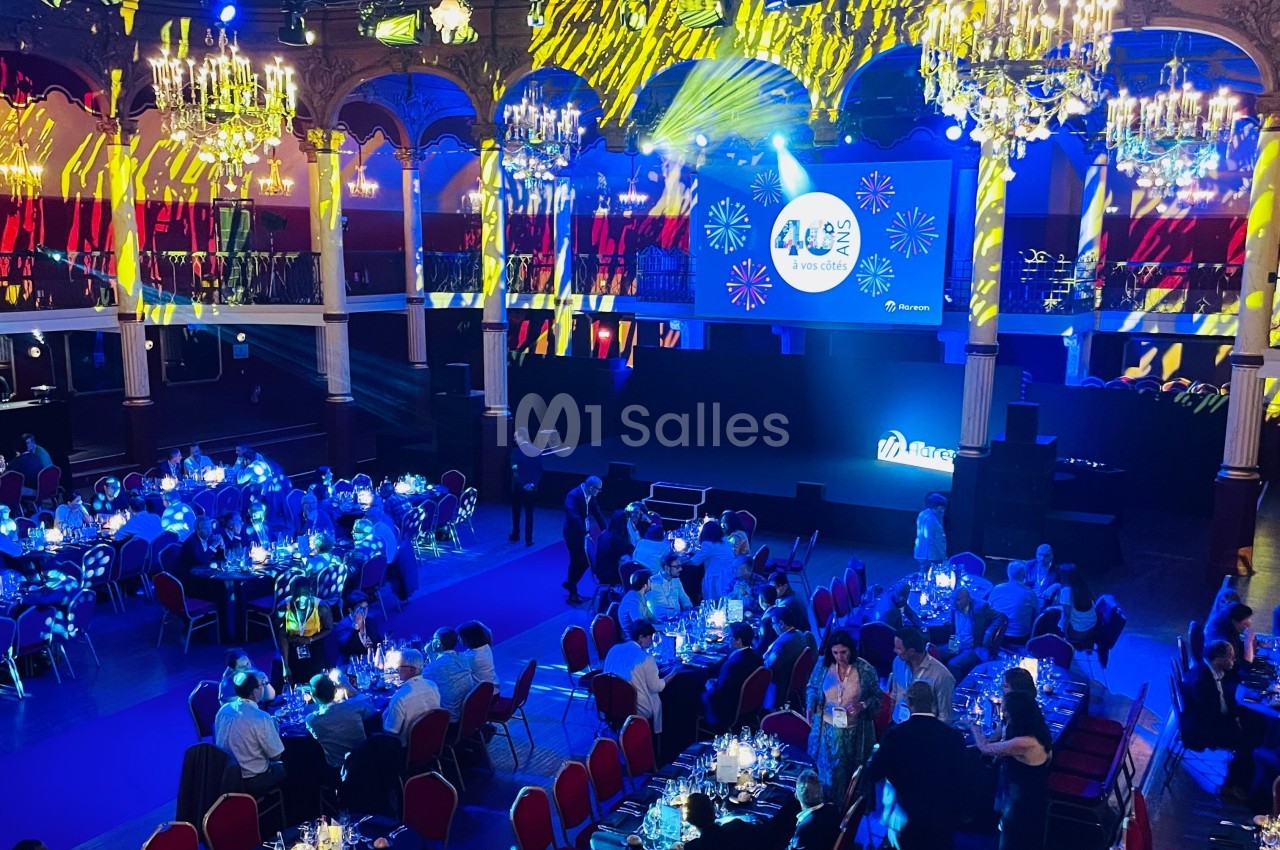 Salle de réception illuminée avec des chandeliers, des projections lumineuses et des invités assis à des tables dressées.