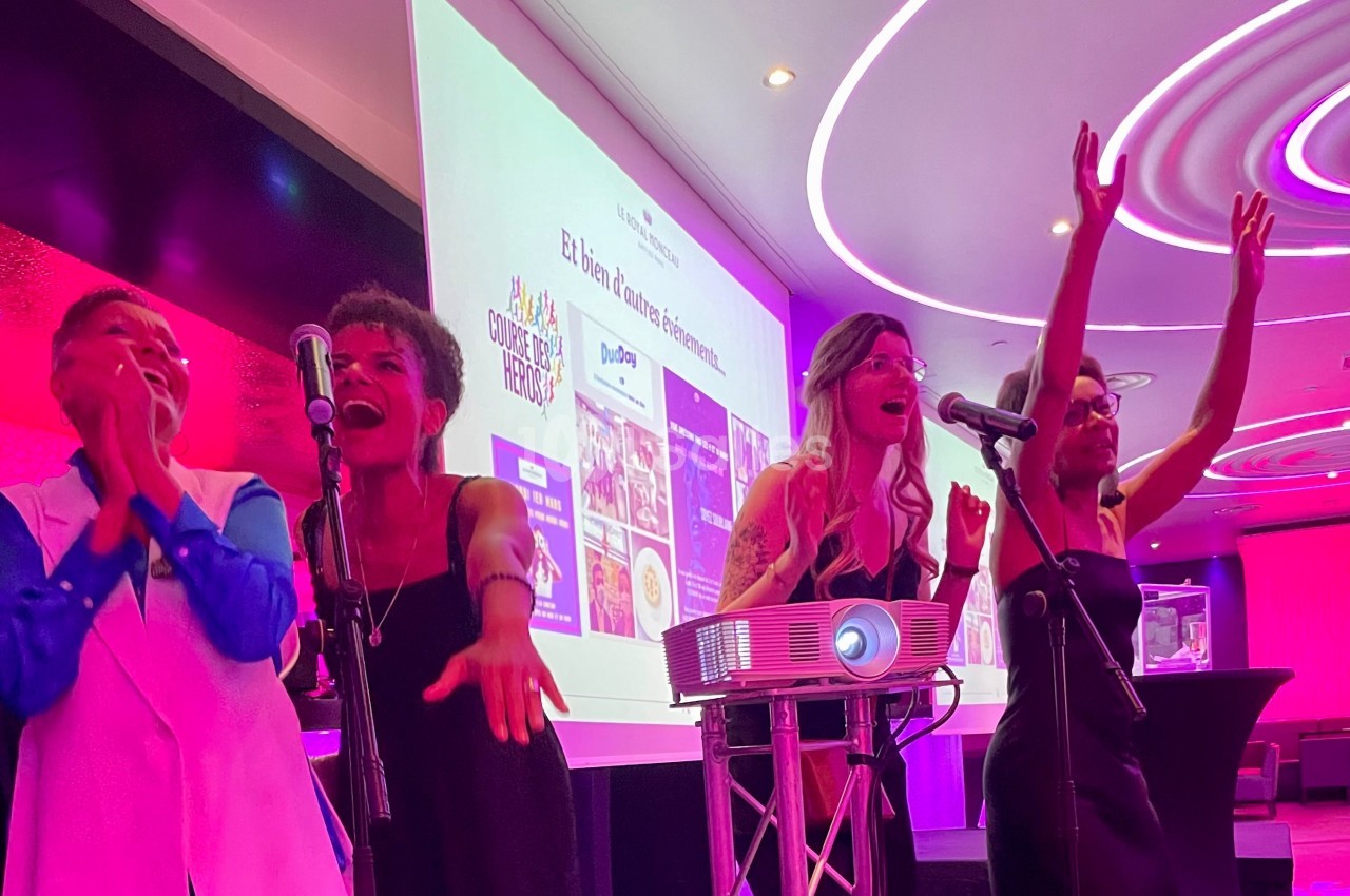 Quatre personnes chantent sur scène avec des micros, dans une salle éclairée par des lumières roses et violettes.