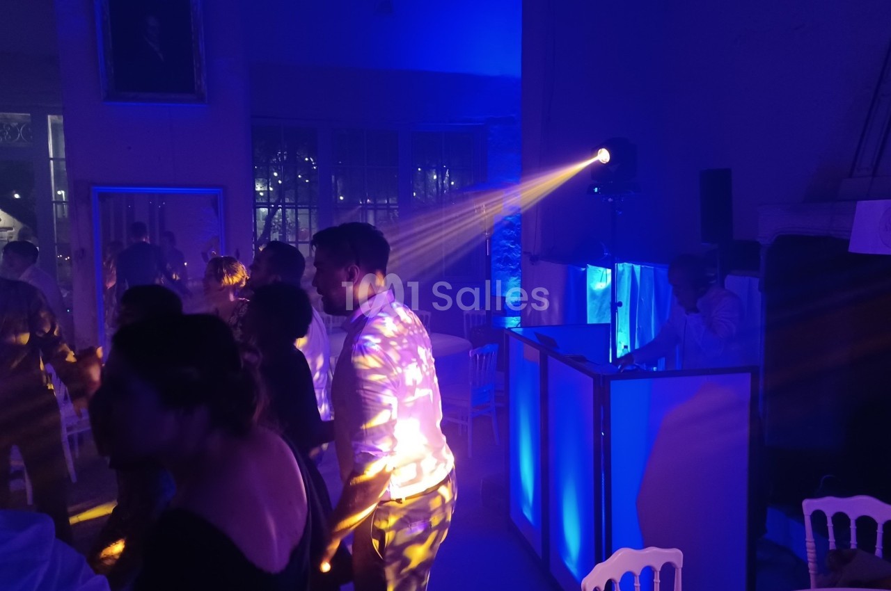 Personnes dansant dans une salle faiblement éclairée avec des lumières colorées et un DJ en arrière-plan.