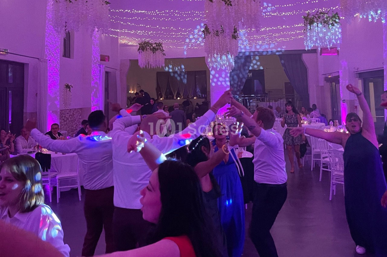 Des invités dansent joyeusement sous des lumières colorées dans une salle décorée pour une fête ou un mariage.