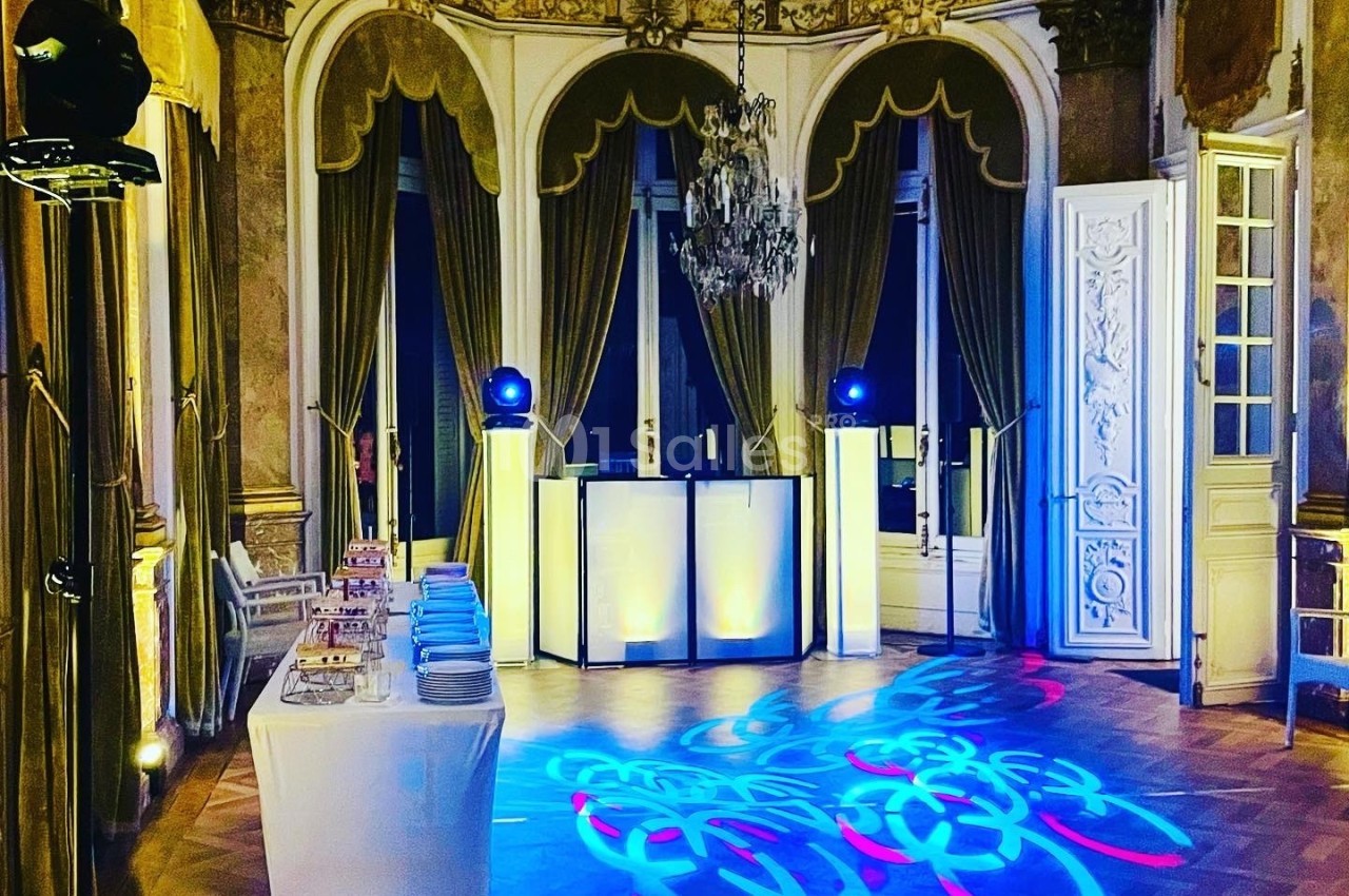 Salle élégante avec parquet, lustre, rideaux dorés, table dressée et éclairages colorés projetant des motifs au sol.