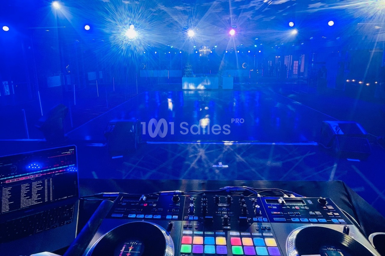 Vue depuis une table de mixage DJ éclairée, face à une piste de danse vide sous des lumières bleues.