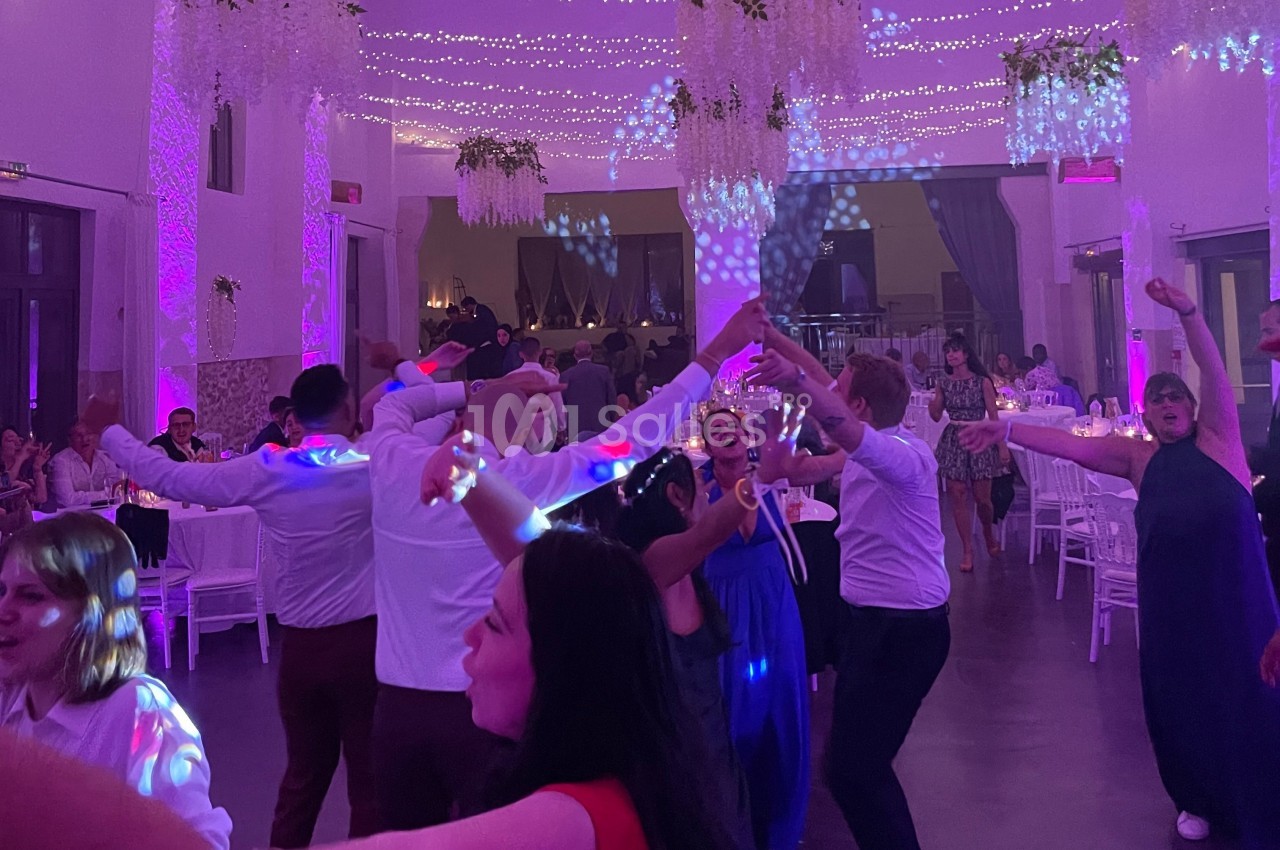 Des invités dansent joyeusement sous des lumières colorées dans une salle décorée pour une fête ou un mariage.