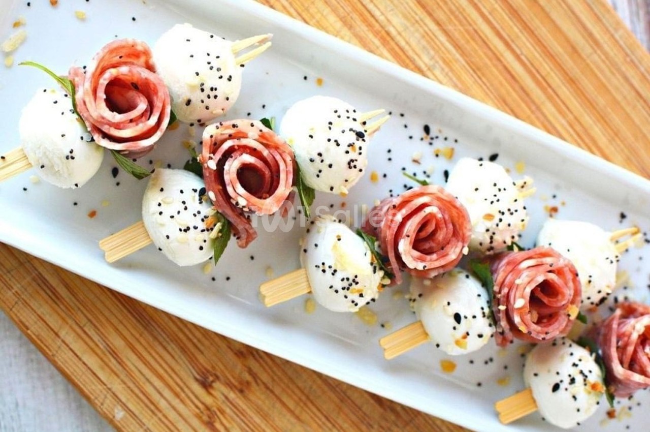 Brochettes apéritives avec mozzarella, tranches de charcuterie en forme de rose, graines et herbes sur un plateau.