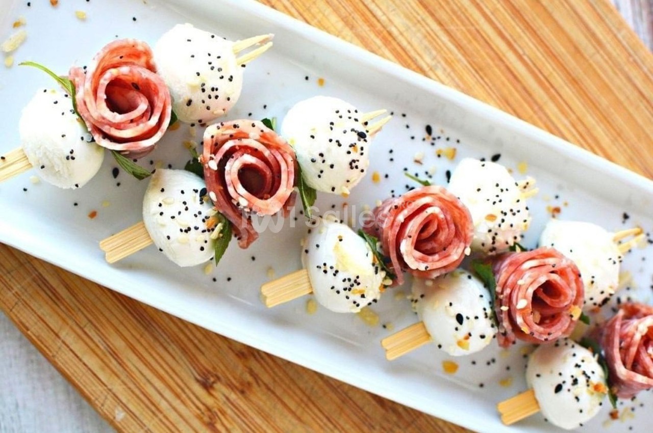 Brochettes apéritives avec mozzarella, tranches de charcuterie en forme de rose, graines et herbes sur un plateau.