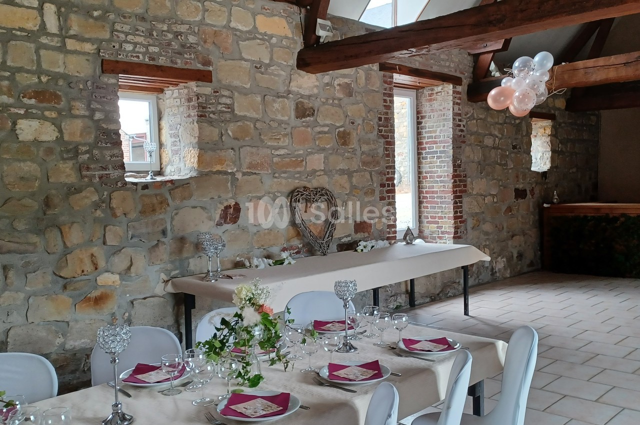 Salle en pierre avec tables dressées pour un événement, décorée de fleurs, bougies et ballons suspendus.