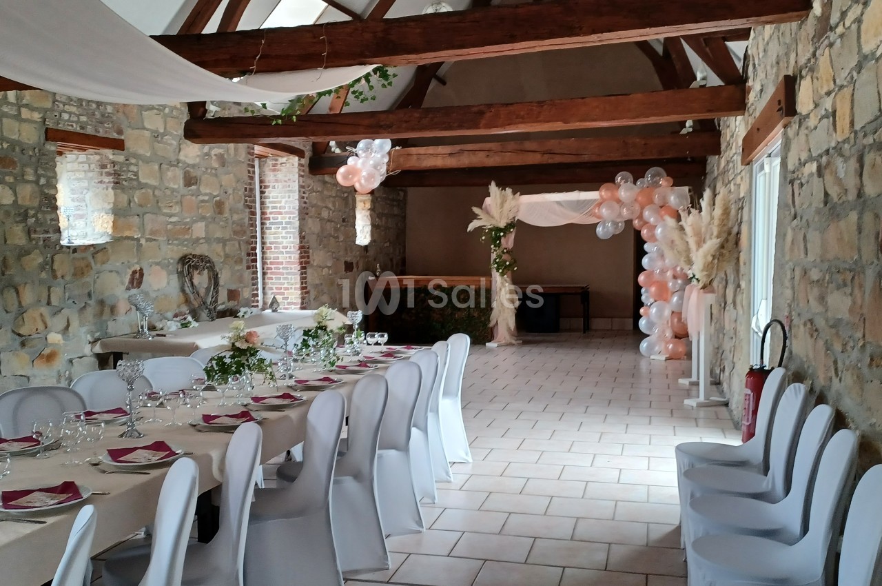 Salle de réception en pierre avec tables dressées, chaises blanches et décoration florale et ballons.