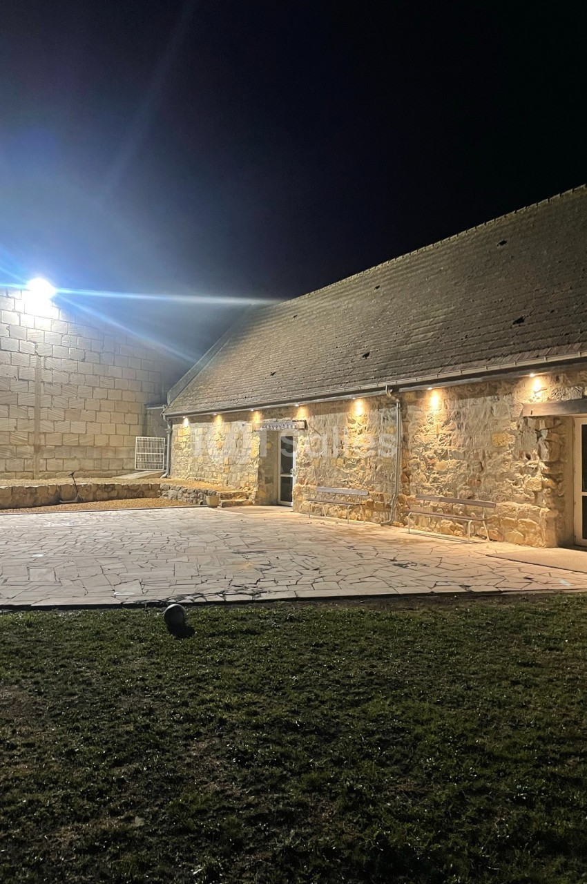 Cour en pierre éclairée la nuit, bordée par un bâtiment en pierre avec des lumières murales et un espace gazonné au premier…