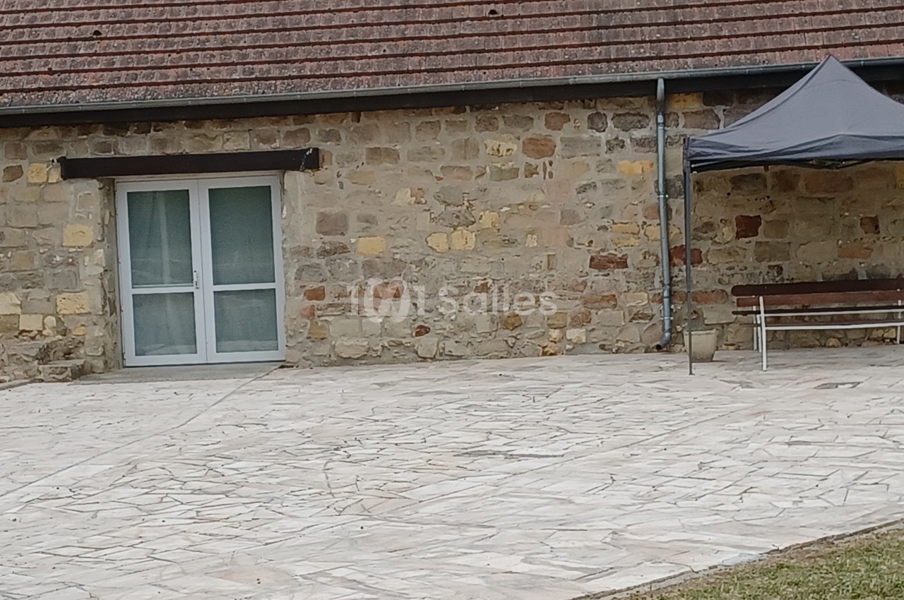 Mur en pierre avec une porte vitrée, une terrasse pavée, un banc en bois et un petit abri gris.