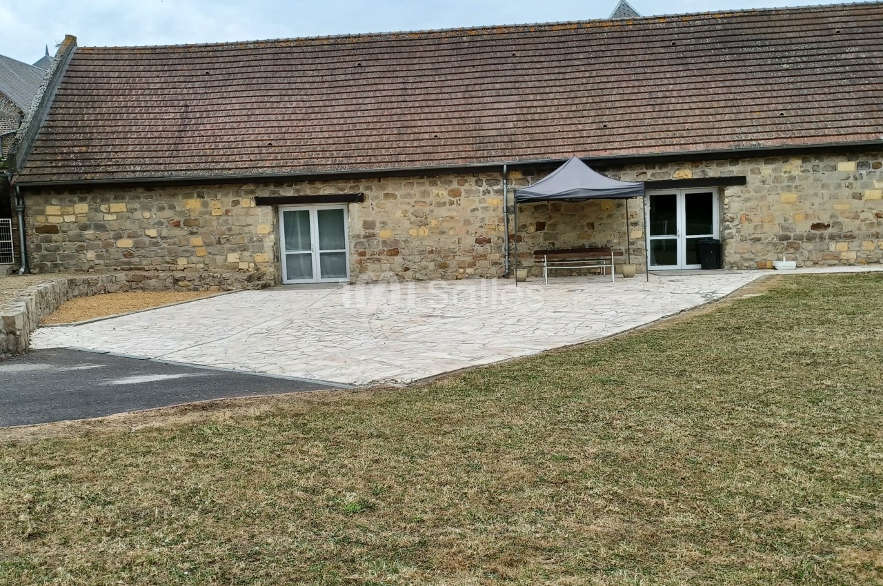 Bâtiment en pierre avec toit en tuiles, terrasse pavée, petite tente et pelouse au premier plan.