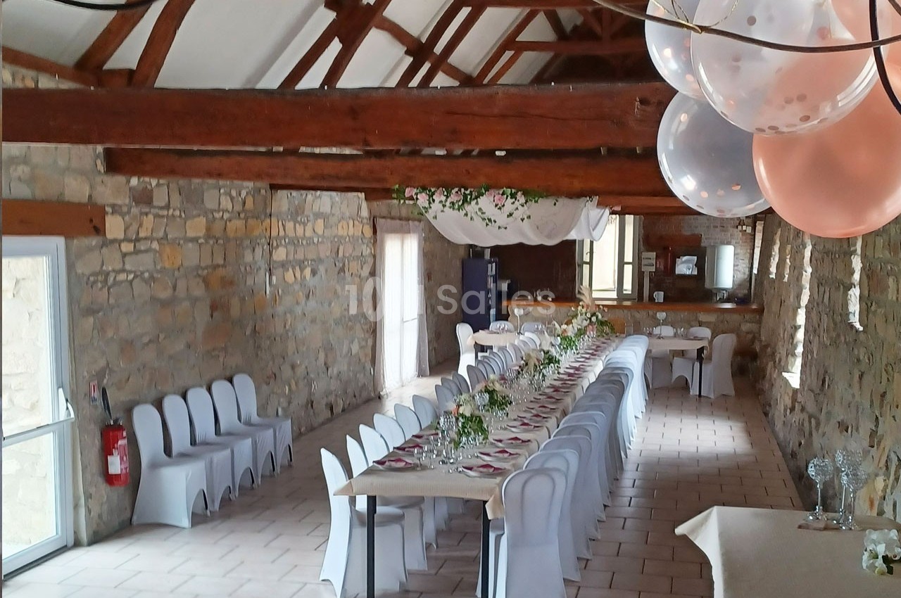 Salle en pierre avec une longue table dressée pour un événement, décorée de fleurs et de ballons suspendus.