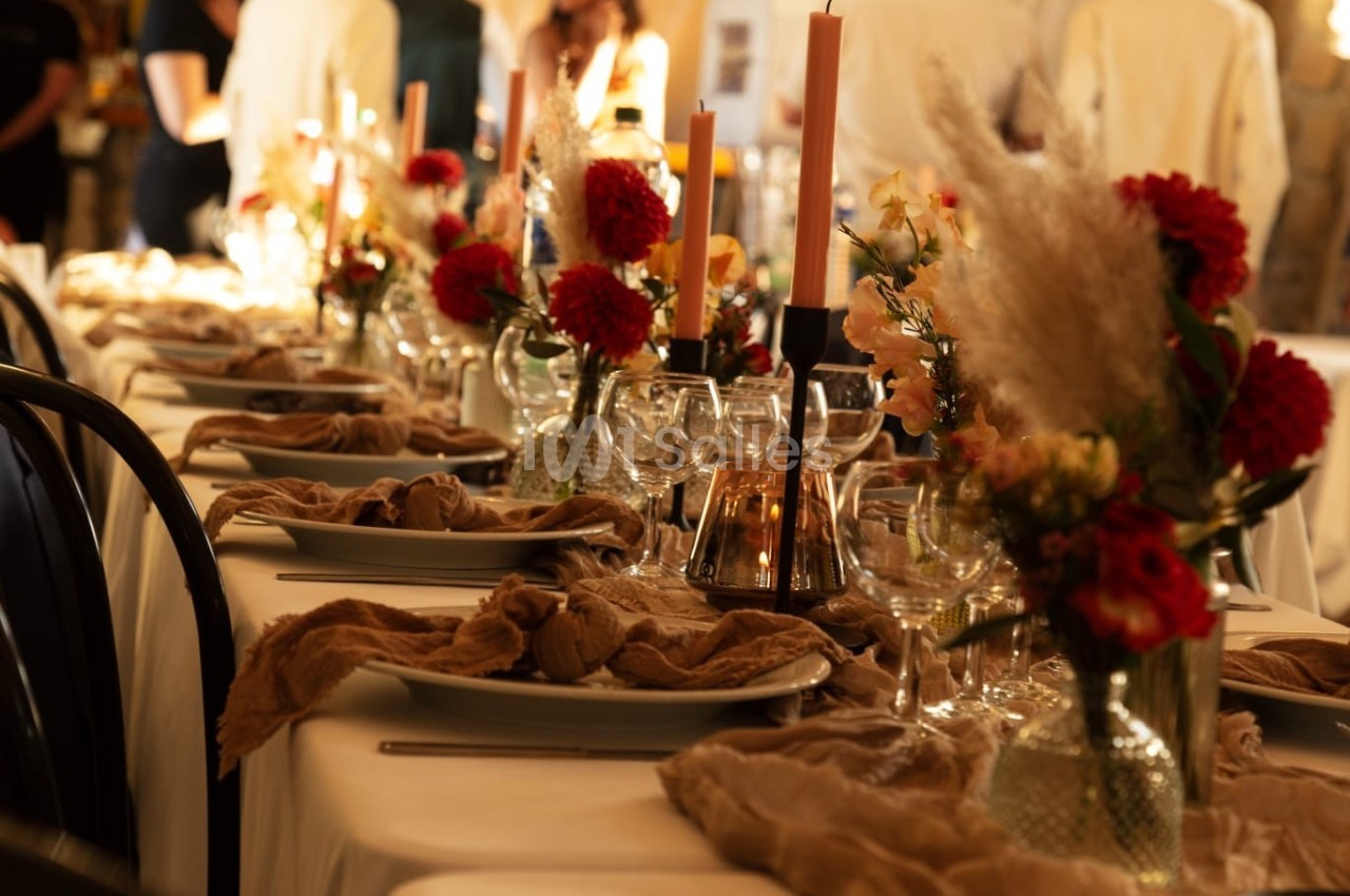 Table décorée avec des fleurs rouges et orangées, bougies allumées et serviettes en tissu beige, ambiance chaleureuse.