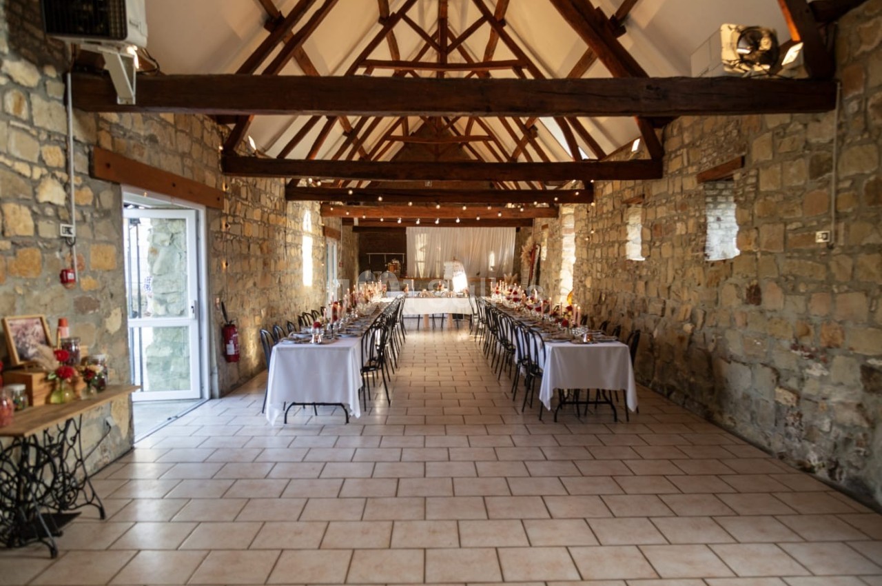 Salle de réception en pierre avec poutres apparentes, tables dressées pour un événement et lumière naturelle.