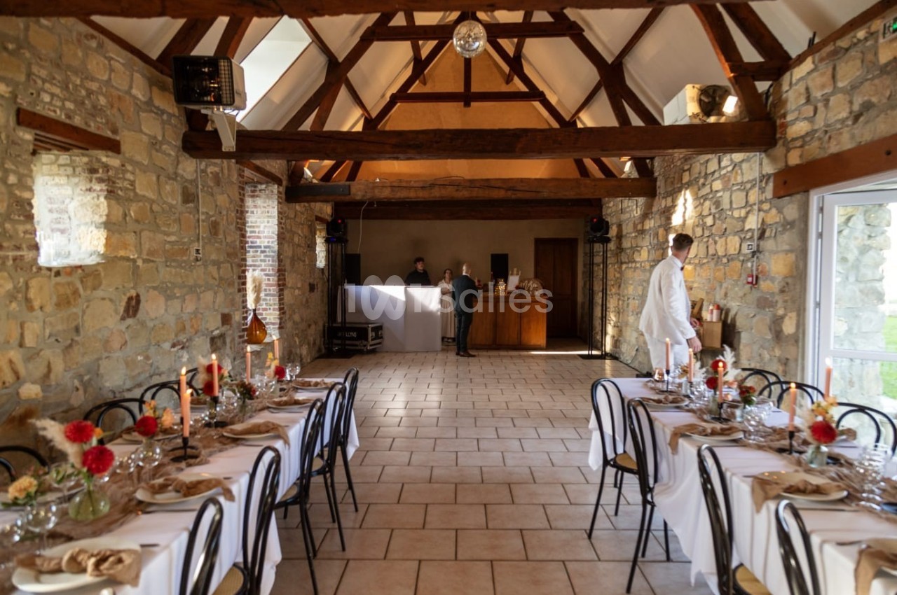 Salle en pierre avec tables dressées pour un repas, décorée de fleurs et bougies, avec un bar en arrière-plan.