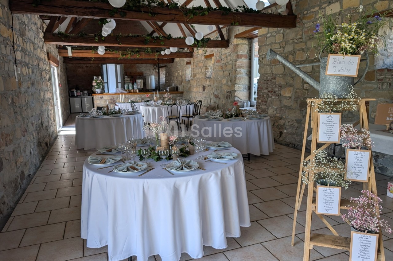 Salle de réception décorée pour un mariage, avec tables rondes dressées et guirlandes végétales suspendues.