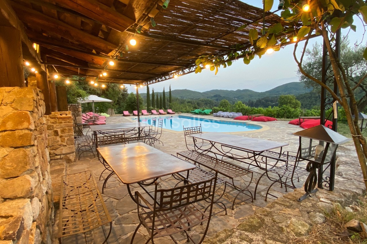 Terrasse en pierre avec tables en fer forgé, éclairée par des guirlandes lumineuses, donnant sur une piscine et un paysage…