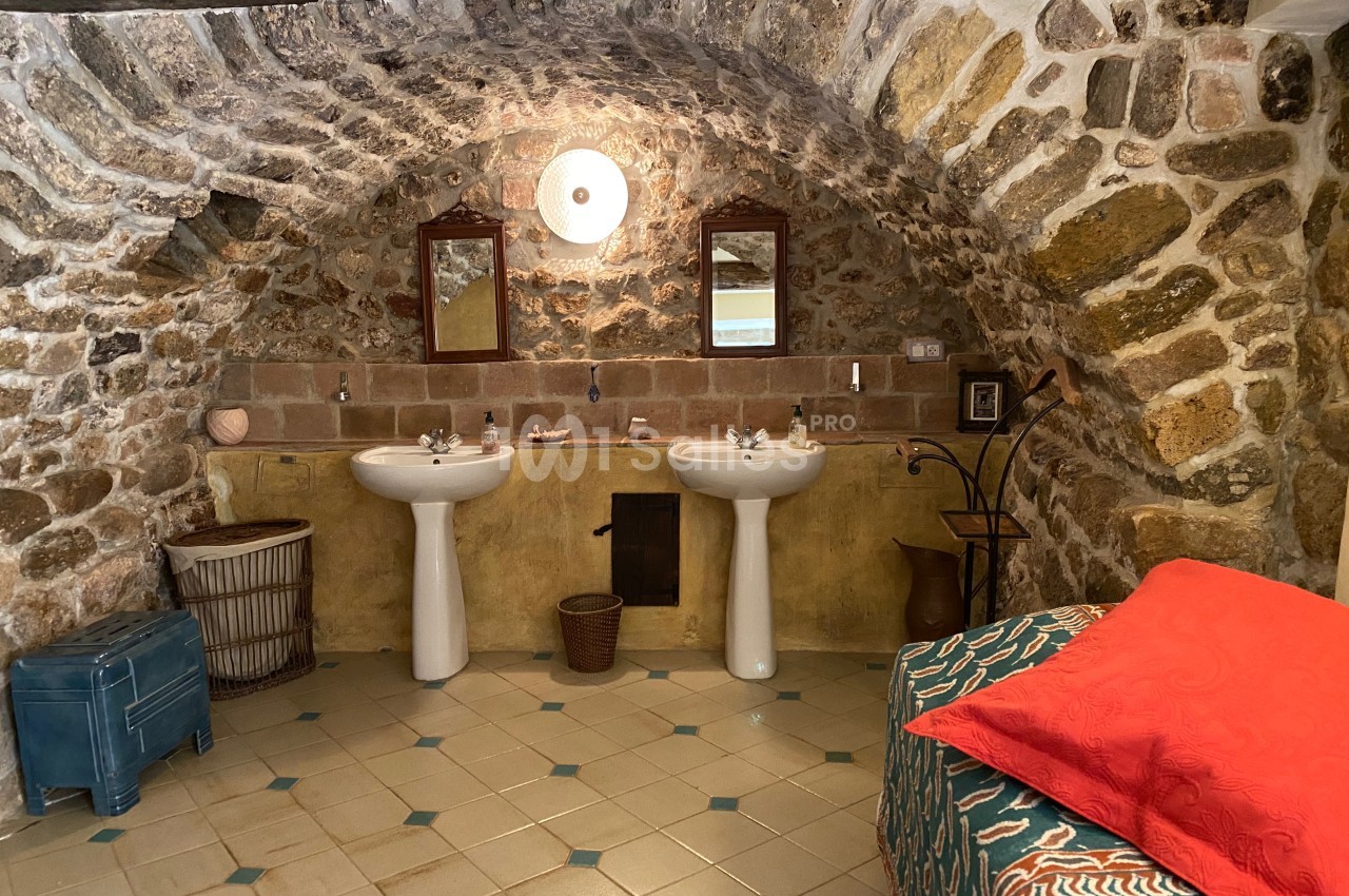 Salle de bain rustique avec murs en pierre voûtés, deux lavabos, un lit avec couvre-lit rouge et un sol carrelé.