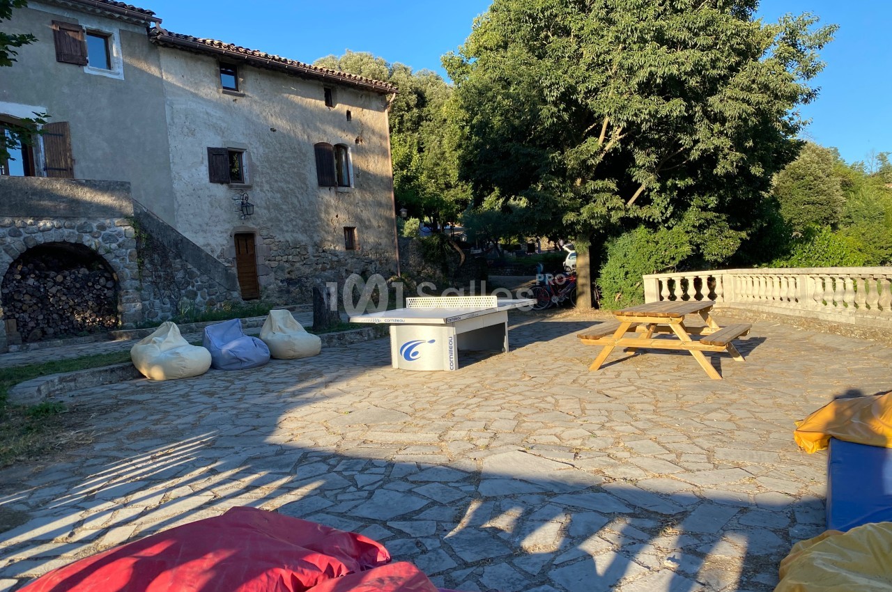 Cour ensoleillée avec table de ping-pong, bancs en bois, poufs colorés et maison en pierre entourée de verdure.