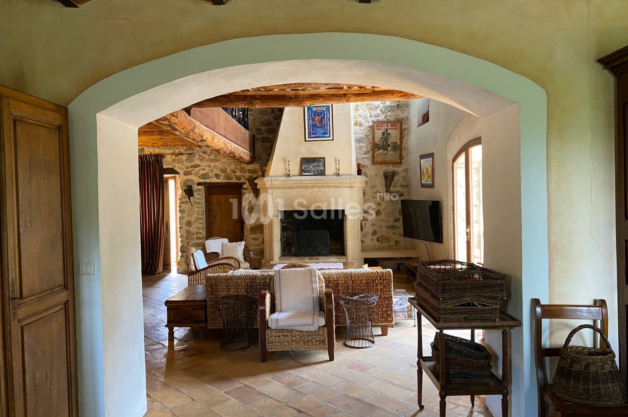 Salon rustique avec cheminée en pierre, poutres apparentes, mobilier en osier et sol en terre cuite.