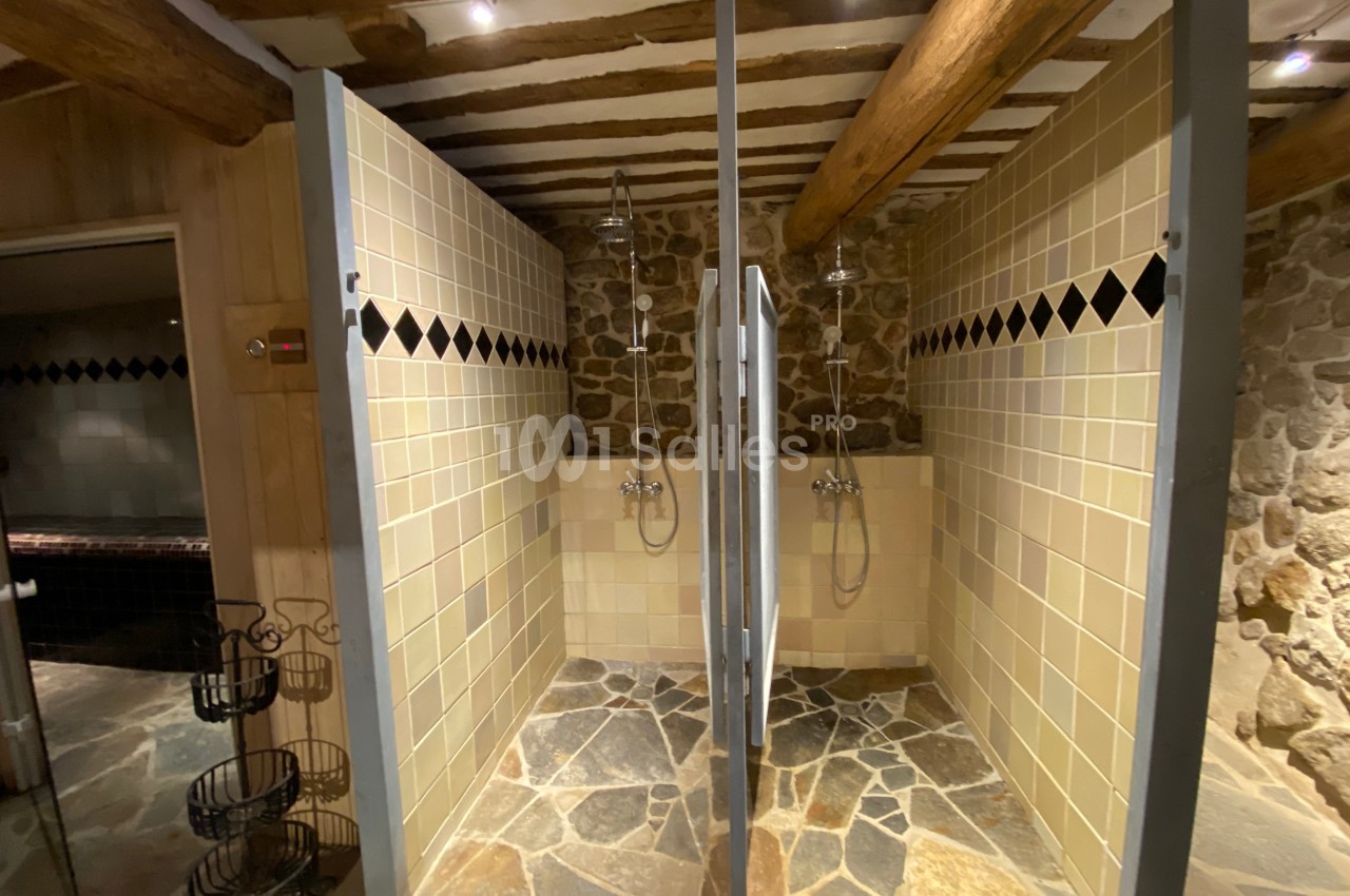 Douches en cabine séparées par des cloisons, avec murs en pierre et carrelage beige sous un plafond en poutres apparentes.