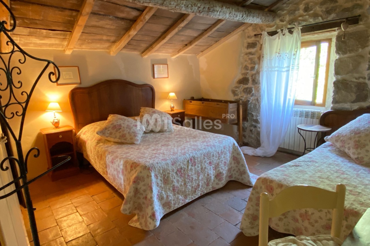 Chambre rustique avec lit double, lit simple, murs en pierre, plafond en bois et lumière naturelle provenant d'une fenêtre.