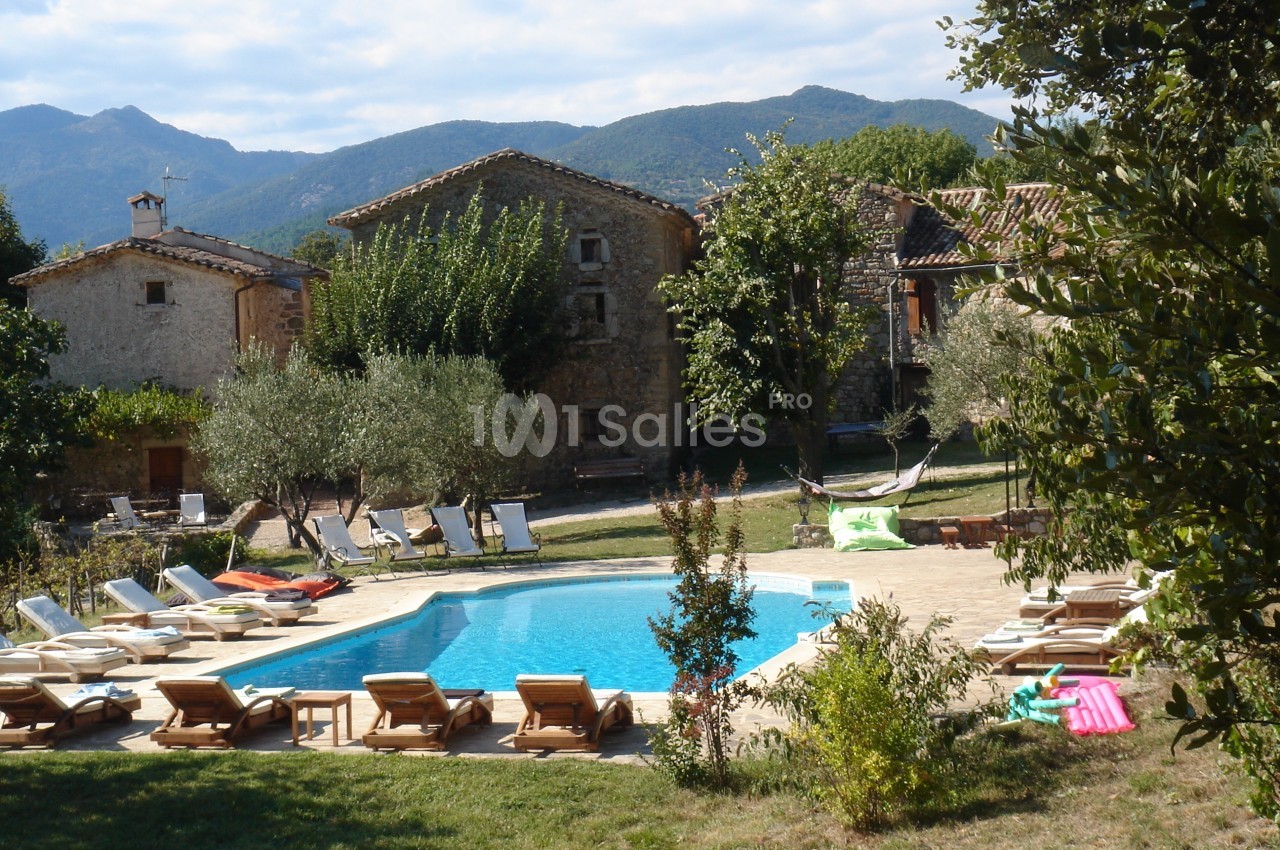 Piscine extérieure entourée de chaises longues, arbres et maisons en pierre, avec des montagnes en arrière-plan.