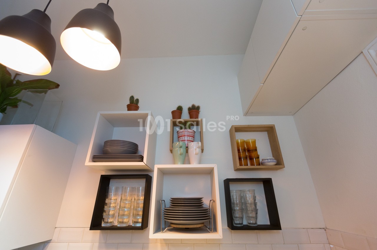 Étagères murales avec vaisselle, verres et plantes en pot dans une cuisine moderne éclairée par des suspensions.