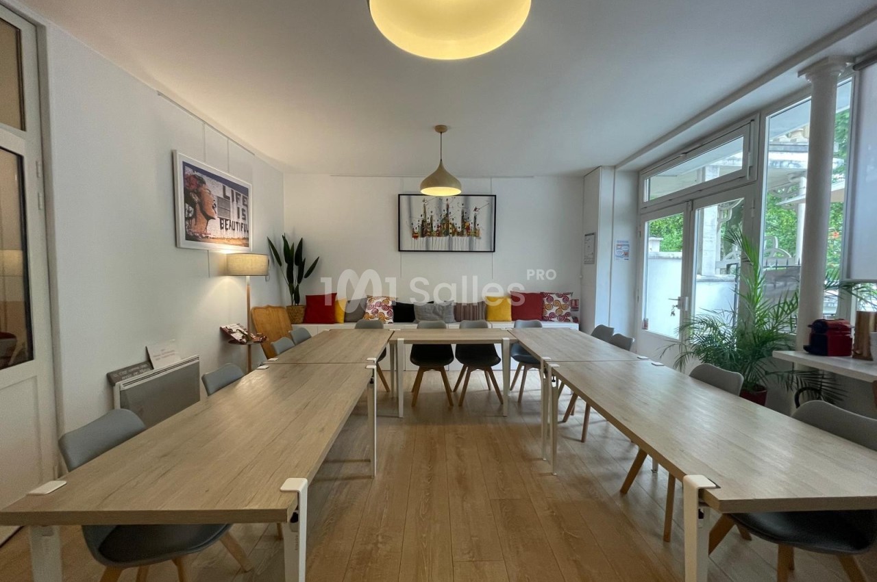Salle lumineuse avec de grandes tables en bois, chaises grises, canapés colorés et décoration murale moderne.