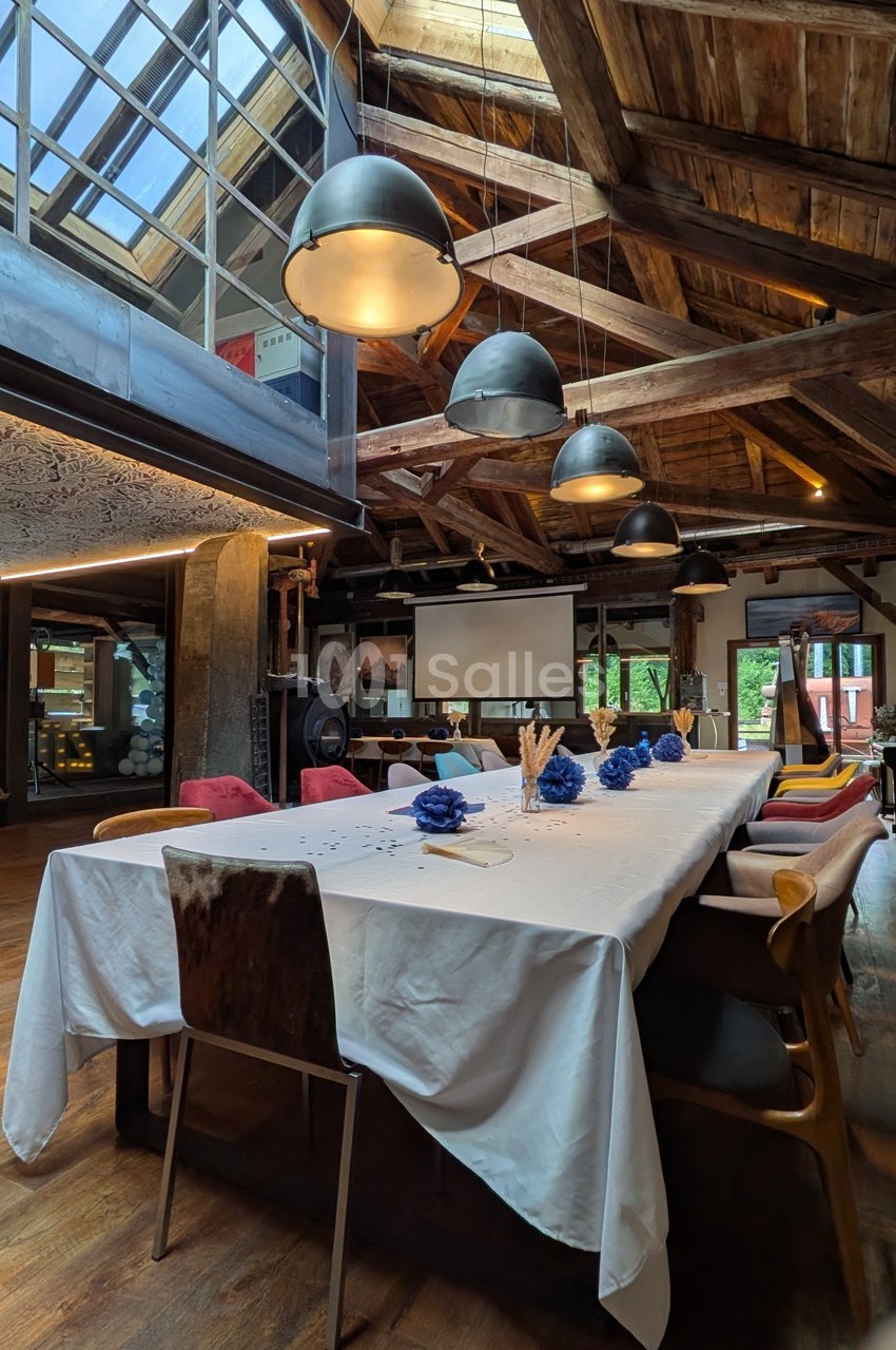 Salle de réunion chaleureuse avec une grande table dressée, des chaises variées et un plafond en bois apparent.