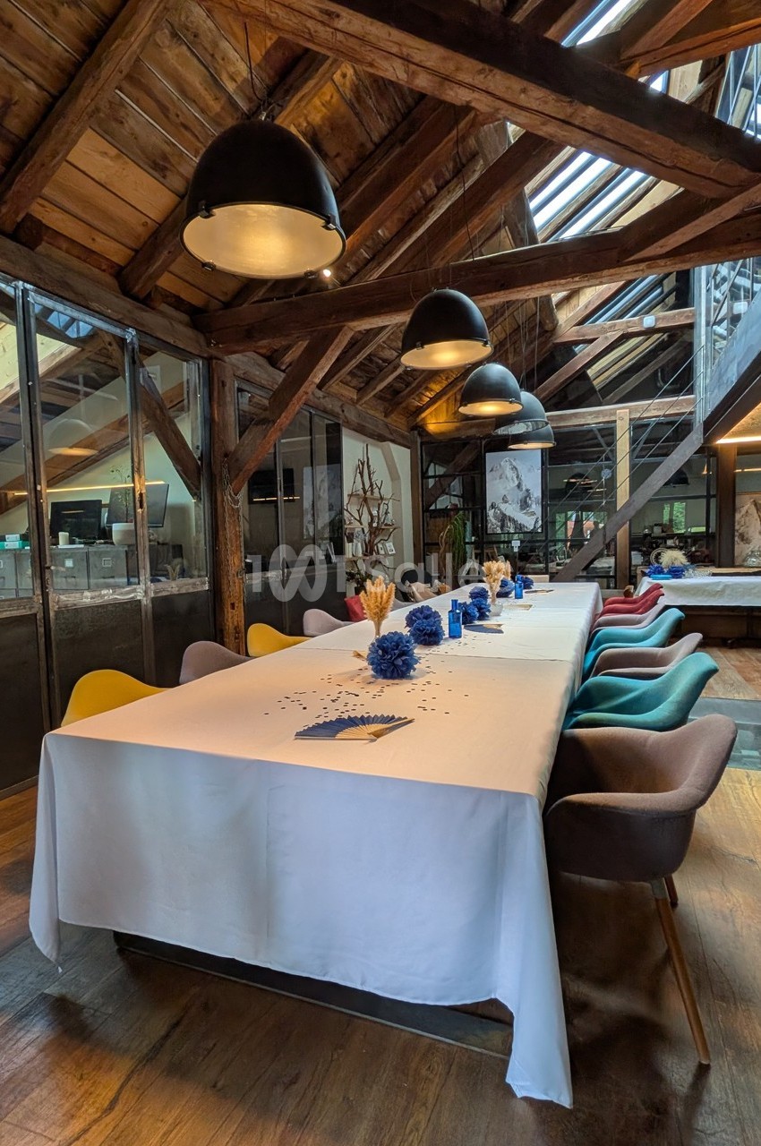 Grande table décorée de fleurs bleues dans une salle lumineuse avec charpente en bois et chaises colorées.
