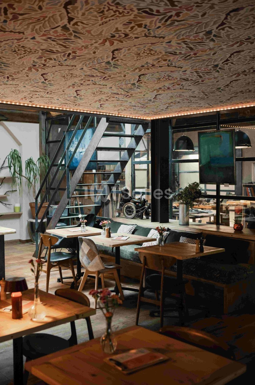 Salle de restaurant moderne avec tables en bois, banquettes, escalier métallique et plafond décoré.