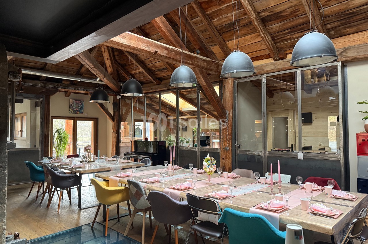 Salle à manger lumineuse avec grande table dressée, chaises colorées et plafond en bois avec poutres apparentes.