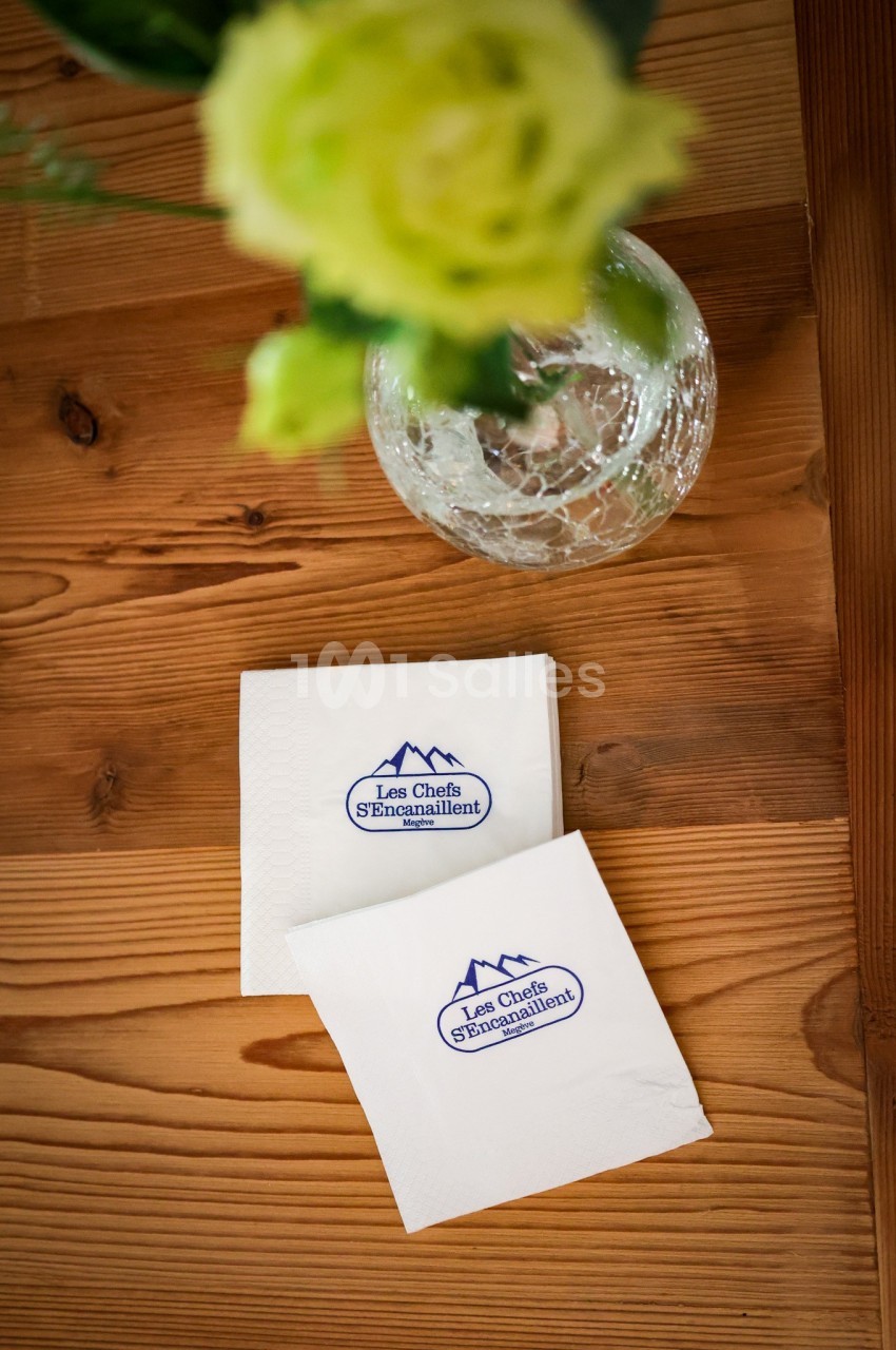 Deux serviettes blanches avec un logo bleu posées sur une table en bois, près d'un vase contenant des fleurs vertes.