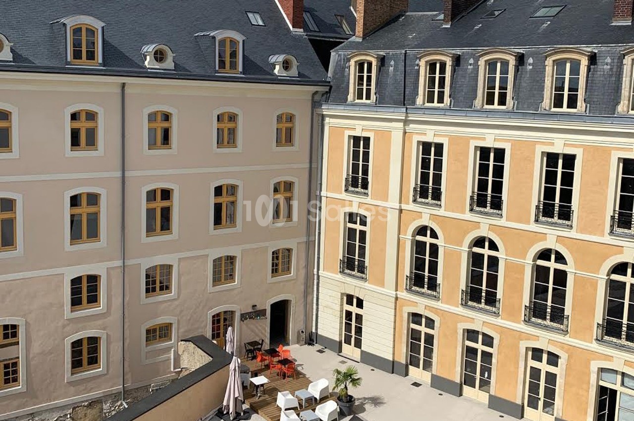 Cour intérieure avec terrasse aménagée, entourée de bâtiments anciens aux façades beige et ocre, fenêtres à volets.