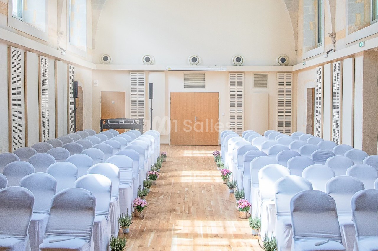 Salle lumineuse avec des rangées de chaises blanches alignées, décorée de plantes et fleurs le long de l'allée centrale.
