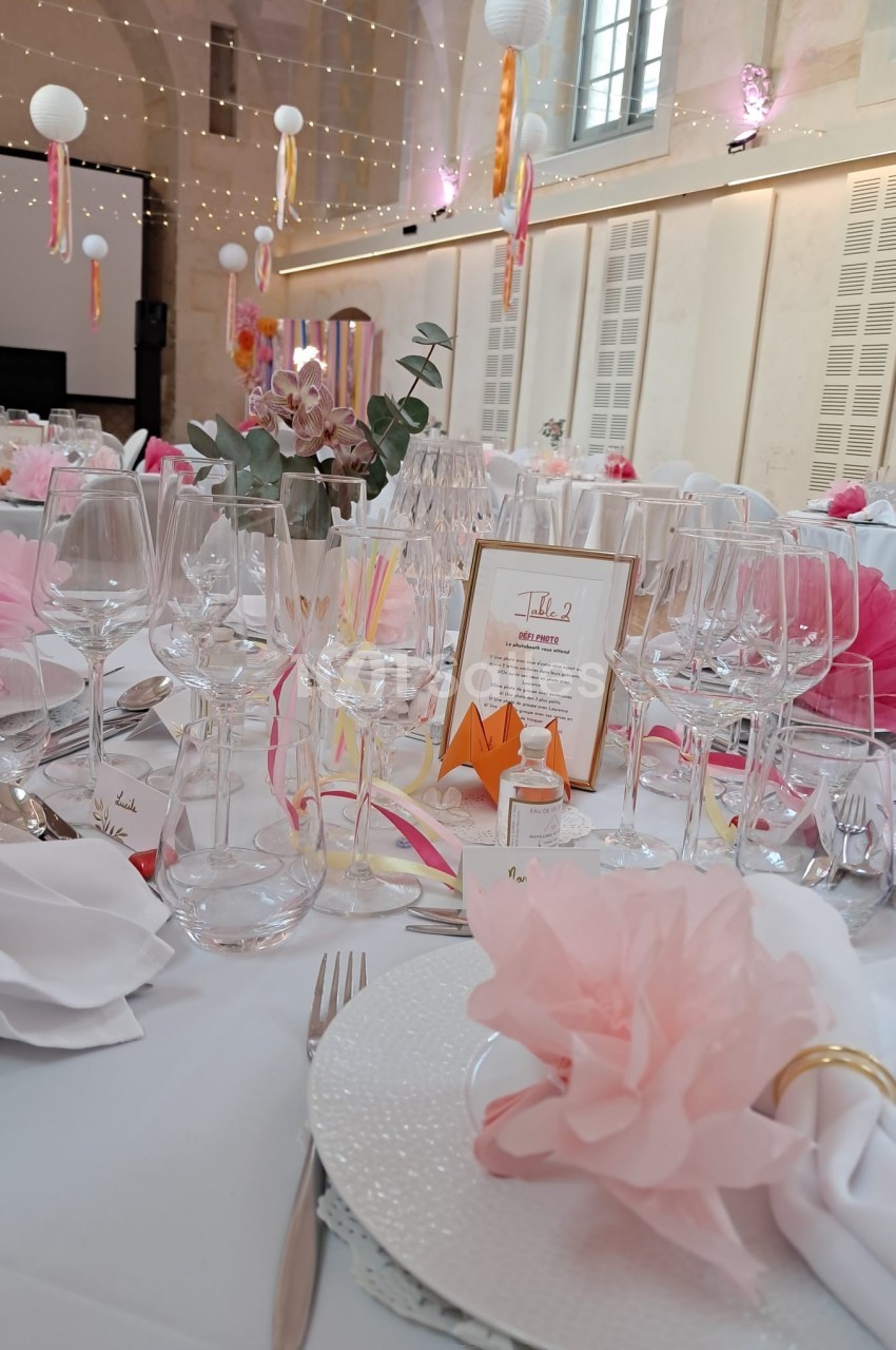 Table décorée pour un mariage avec fleurs roses, verres, menus et guirlandes lumineuses en arrière-plan.