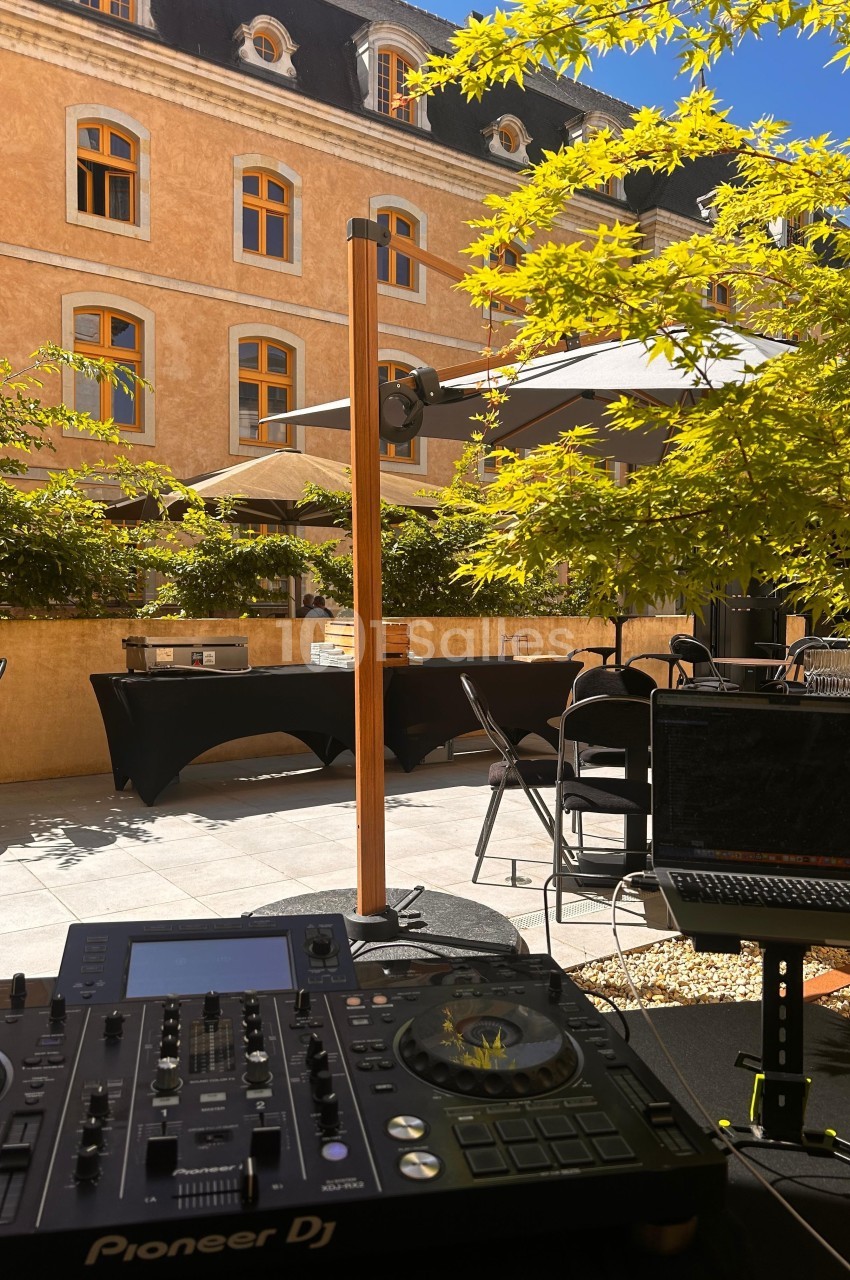 Console DJ en premier plan sur une terrasse ensoleillée, avec tables, parasols et bâtiment en arrière-plan.