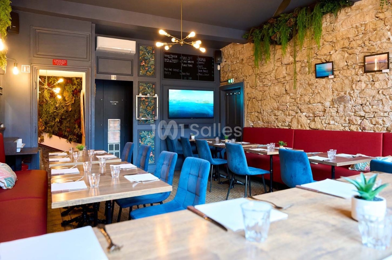 Salle de restaurant lumineuse avec murs en pierre, tables dressées, chaises colorées et décoration végétale.