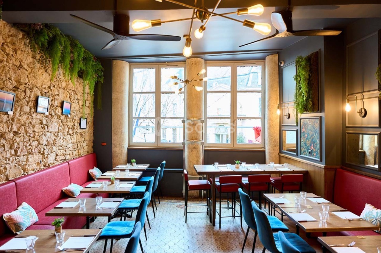 Salle de restaurant lumineuse avec murs en pierre, banquettes rouges, chaises bleues et grandes fenêtres.