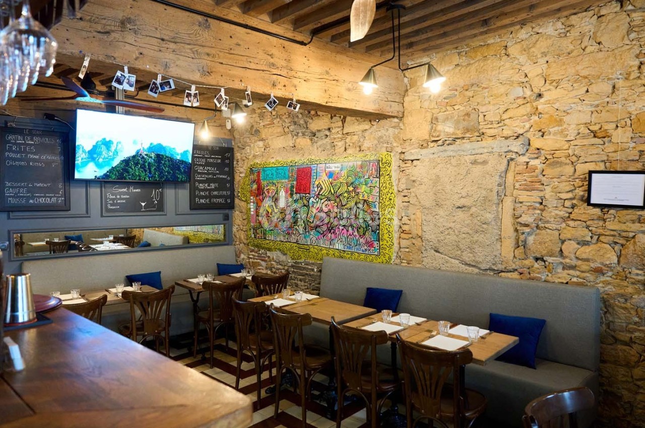 Salle de restaurant avec murs en pierre, tables dressées, banquettes grises et décorations murales colorées.