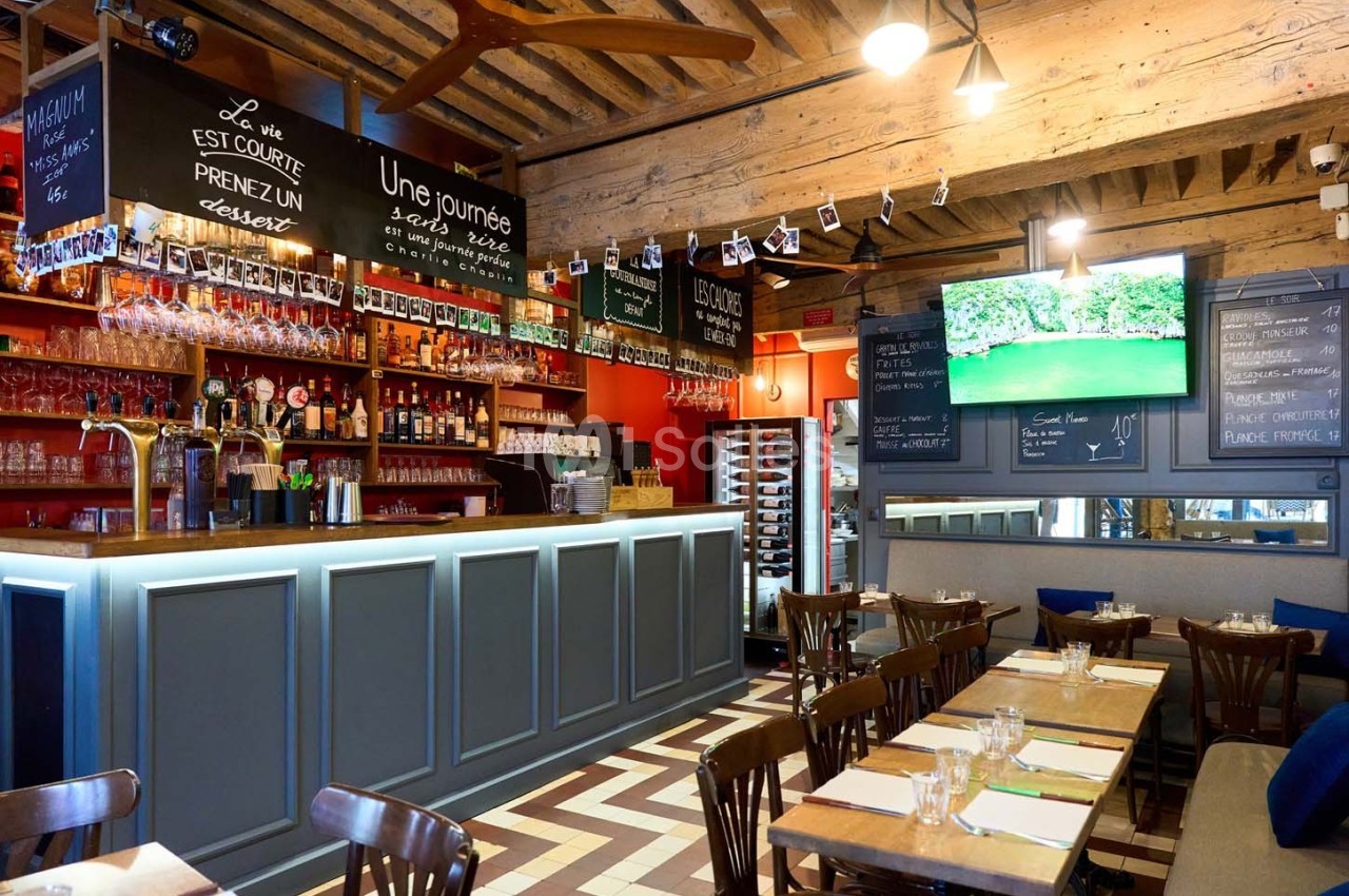 Intérieur d'un restaurant avec bar en bois, étagères de bouteilles, tables dressées et écran affichant un paysage.