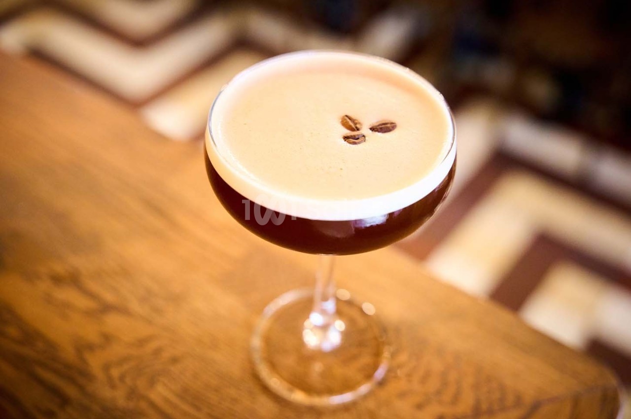 Verre à cocktail rempli d'une boisson mousseuse, décorée de trois grains de café, posé sur une table en bois.